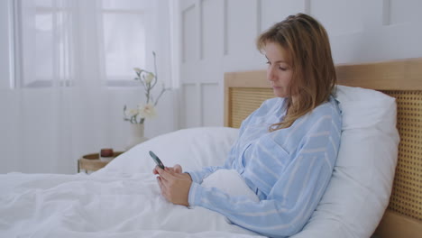 Attractive-young-beautiful-girl-chatting-with-her-friends--while-sitting-on-her-bedroom.-Young-woman-lying-in-bed-and-holding-smartphone